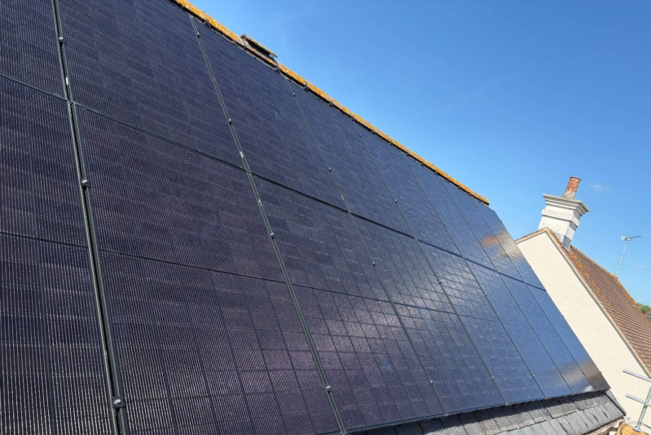 Two rows of all-black solar panels mounted in portrait orientation on a grey tiled pitched roof, with bird mesh fitted along the eaves, photographed from the scaffold at a Wiltshire installation.